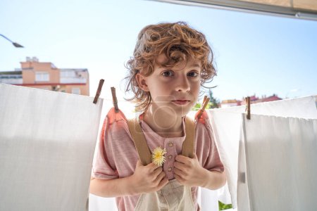 Foto de Adorable niño preadolescente con flor amarilla que se divierte mientras hace la colada y se cuelga en el tendedero cerca de sábanas blancas con pinzas de ropa en el balcón en casa - Imagen libre de derechos