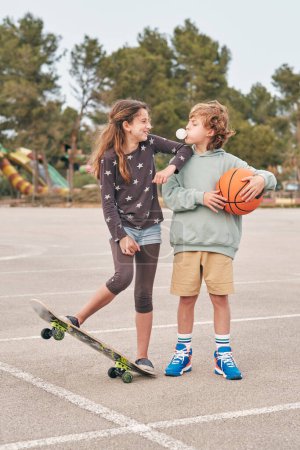 Téléchargez les photos : Positif adolescent garçon et fille debout avec skateboard et basket en ville et souffler chewing-gum tout en s'amusant et en se regardant - en image libre de droit