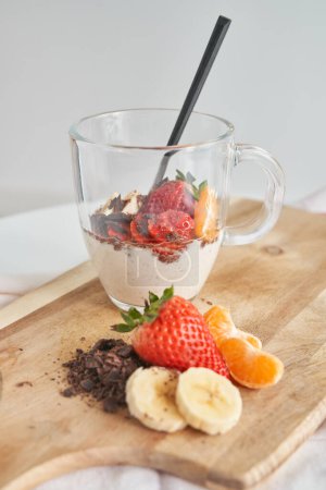 Téléchargez les photos : Dessert à la fraise fraîche avec chocolat dans une tasse en verre avec cuillère et tranches de mandarine de banane placées sur une planche à découper en bois - en image libre de droit