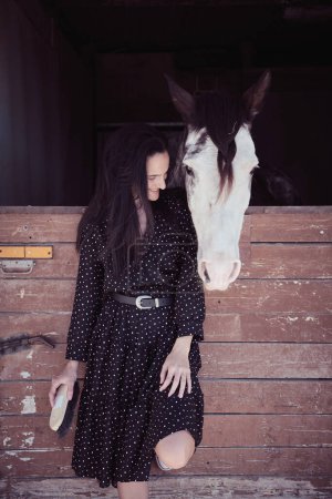 Foto de Contenida propietaria femenina con cepillo en vestido negro mirando obediente caballo blanco de pie en un establo de madera en el campo - Imagen libre de derechos