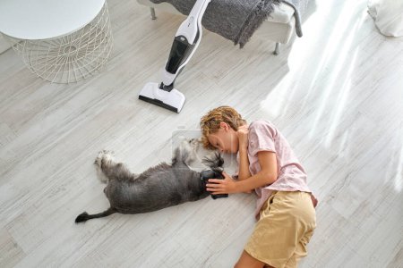 Téléchargez les photos : De dessus garçon en vêtements décontractés caressant chien Schnauzer tout en étant couché sur le sol près de l'aspirateur pendant la routine du ménage - en image libre de droit