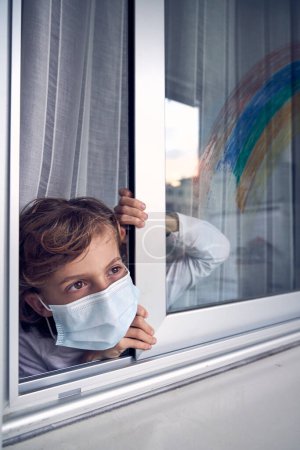 Foto de Niño preadolescente con máscara médica protectora de pie cerca de la ventana y mirando hacia otro lado mientras se queda en casa durante la epidemia de coronavirus - Imagen libre de derechos