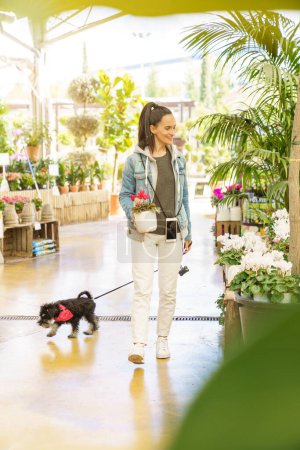 Téléchargez les photos : Corps complet de femme hispanique positive propriétaire avec chien marchant dans une boutique florale moderne spacieuse avec diverses plantes à fleurs en pot - en image libre de droit
