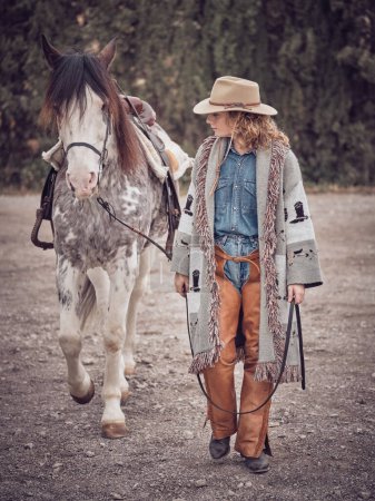 Foto de Cuerpo completo de hembra en sombrero y ropa occidental caminando cerca de caballo obediente con brida en el campo con árboles verdes - Imagen libre de derechos