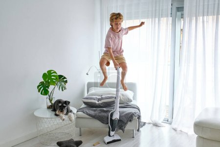 Photo for Barefoot boy leaping from chair while tidying floor near table with Schnauzer dog while doing housework in light living room - Royalty Free Image