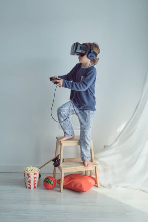 Foto de Vista lateral del niño preadolescente de cuerpo completo en gafas VR y auriculares parados en la escalera de taburete cerca de palomitas de maíz y jugando con joystick - Imagen libre de derechos