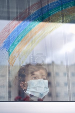 Photo for Through glass view of kid in protective mask standing near window with curtains while looking away during quarantine period at home - Royalty Free Image