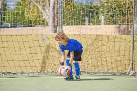 Photo for Full length of preteen goalkeeper in activewear leaning forward while standing at goal with ball in hands during soccer training on sports ground - Royalty Free Image
