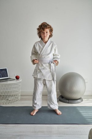 Photo for Full length of barefoot child in kimono and white belt standing on sports mat in pose and looking at camera while practicing judo at home - Royalty Free Image