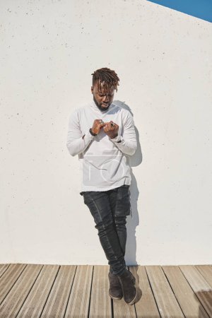 Photo for Full body of bearded African American with dreadlocks in trendy outfit standing with crossed legs and leaning on wall while looking at hands - Royalty Free Image