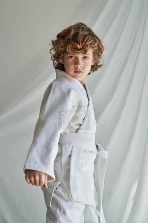 Foto de Niño preadolescente sin emociones con el pelo rubio rizado en kimono blanco de pie cerca de la cortina con el puño cerrado y mirando a la cámara mientras practica judo - Imagen libre de derechos