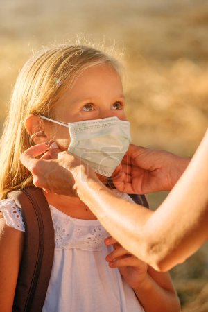 Photo for Crop person putting protective mask on adorable preteen child with backpack while standing in nature during coronavirus pandemic - Royalty Free Image