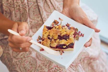 Téléchargez les photos : Femme anonyme en robe assise sur une chaise et mangeant une tarte aux baies appétissante décorée de pétales de fleurs et servie sur une assiette blanche à l'aide d'une fourchette en bois jetable - en image libre de droit
