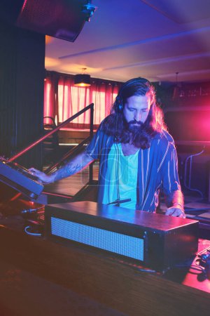 Photo for Concentrated bearded DJ with headphones playing music on modern musical equipment at table while working in nightclub with dim illumination - Royalty Free Image