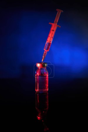 Foto de Jeringa insertada en frasco con vacuna contra el coronavirus en luz roja sobre superficie reflectante sobre fondo azul - Imagen libre de derechos
