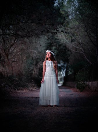 Photo for Full body of girl wearing white dress standing on pathway near green trees and dark bushes - Royalty Free Image