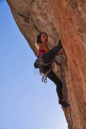 Photo for From below of female alpinist wearing safety harness sportswear and boots climbing steep cliff using dynamic rope carabines and chalkbag and looking away - Royalty Free Image