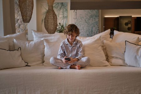 Téléchargez les photos : Corps complet d'un garçon aux cheveux bouclés avec les jambes croisées pieds nus en pyjama assis sur le canapé et naviguant sur son téléphone portable tout en se reposant dans un salon confortable à la maison - en image libre de droit
