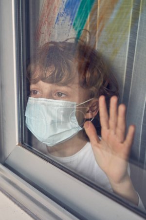 Photo for Adorable boy in protective medical mask looking away through window while touching glass with hand during coronavirus pandemic - Royalty Free Image