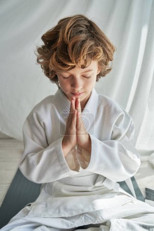 Photo for Concentrated preteen boy with curly hair in white kimono practicing yoga on mat in Lotus pose with closed eyes and namaste gesture near curtain - Royalty Free Image
