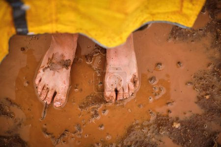 Téléchargez les photos : Vue du dessus d'une personne anonyme pieds nus sans visage en manteau jaune debout dans une flaque de boue - en image libre de droit