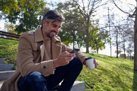 Photo for Low angle of middle aged positive unshaven Hispanic man in beige coat using cellphone and enjoying coffee break while sitting on stone stairs against trees and green hillside - Royalty Free Image