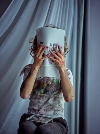 Téléchargez les photos : Enfant créatif méconnaissable en t-shirt teinté de peinture se cachant derrière une feuille de papier avec des empreintes de mains colorées sur fond gris - en image libre de droit