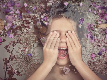 Foto de Vista superior del niño cubriendo los ojos y gritando mientras está acostado en agua transparente con flores de colores en el baño de luz - Imagen libre de derechos