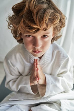 D'en haut de l'enfant aux cheveux bouclés concentré en kimono blanc méditant avec geste namaste tout en pratiquant le yoga à Padmasana et en regardant la caméra