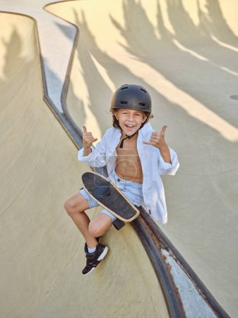 Foto de Desde arriba toda la longitud del joven patinador con longboard gesto shaka y sentado en la rampa en el parque de skate en el día soleado - Imagen libre de derechos