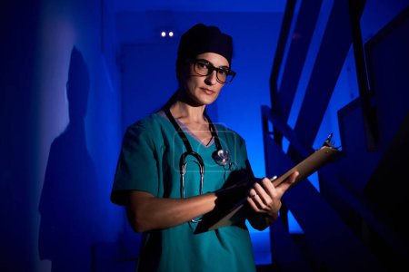 Foto de Médico profesional en uniforme médico con estetoscopio escrito en el portapapeles y mirando a la cámara mientras está de pie en las escaleras en la habitación oscura bajo iluminación azul - Imagen libre de derechos
