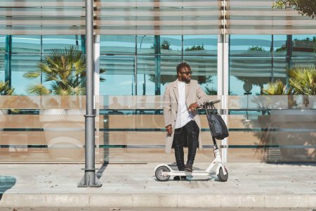 Téléchargez les photos : Vue latérale de l'homme afro-américain avec des lunettes en robe élégante debout sur le trottoir avec trottinette scooter électrique sur la rue de la ville tout en regardant loin - en image libre de droit
