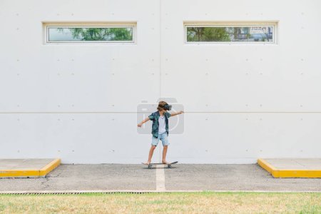 Foto de Vista lateral de un niño irreconocible que experimenta la realidad virtual mientras monta monopatín en el pavimento de la calle de hormigón cerca de la pared - Imagen libre de derechos