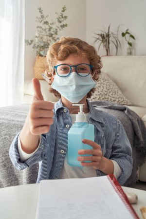 Photo for Child in medical mask and glasses sitting at table with dispenser of antibacterial gel in hand and looking at camera while showing thumb up gesture during COVID epidemic - Royalty Free Image