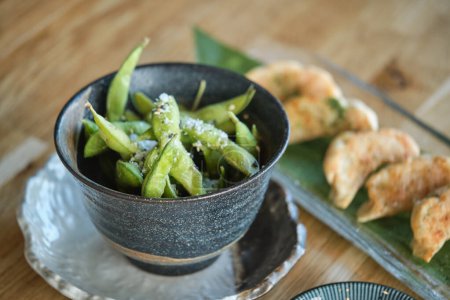Photo for Bowl with green edamame beans served near tasty fried jiaozi dumplings on banana leaf in light restaurant with Asian food - Royalty Free Image