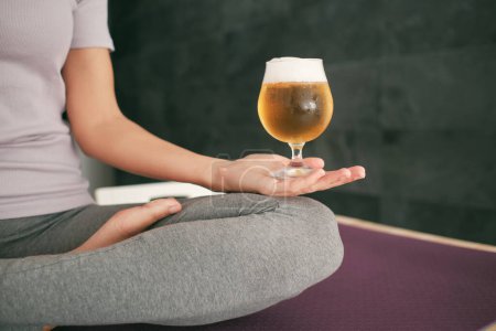 Photo for Crop anonymous barefoot female in activewear meditating with glass of fresh foamy beer while sitting on mat in light room - Royalty Free Image