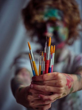 Foto de Enfoque suave del niño anónimo cubierto de pinturas que muestran un conjunto de pinceles en manos sucias mientras está de pie en la sala de luz - Imagen libre de derechos