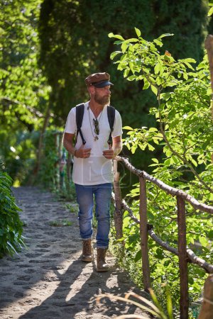 Téléchargez les photos : Corps complet de mâle barbu en vêtements décontractés et chapeau admirant la nature tout en marchant sur une allée pavée dans la forêt - en image libre de droit