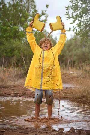 Téléchargez les photos : Pleine longueur de l'enfant en imperméable jaune criant et versant de l'eau sale hors des bottes de gomme tout en se tenant dans la flaque d'eau dans les bois le jour de pluie - en image libre de droit