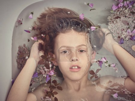 Photo for Top view of cute boy touching hair and looking at camera while lying in clean bathtub with colorful flowers in bathroom - Royalty Free Image