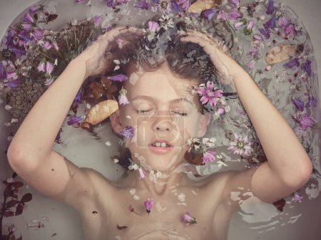 Téléchargez les photos : Vue du dessus du garçon calme avec les yeux fermés couché sous l'eau de la baignoire claire avec de petites fleurs colorées dans la salle de bain légère - en image libre de droit