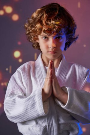 Foto de Niño preadolescente tranquilo en kimono blanco mirando hacia otro lado con las manos dobladas en saludo mientras está de pie en el estudio durante la práctica de judo - Imagen libre de derechos