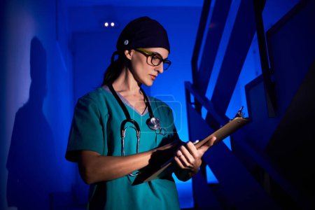 Téléchargez les photos : Femme concentrée en peignoir médical écrivant sur le presse-papiers tout en se tenant sur les escaliers dans l'hôpital sombre sous la lumière néon bleu - en image libre de droit
