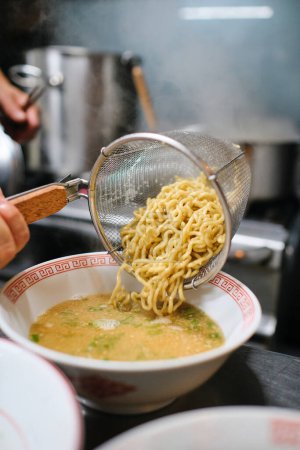 Téléchargez les photos : Chef méconnaissable ajoutant des nouilles du tamis dans un bol avec un délicieux bouillon avec assaisonnement dans le processus de faire de la soupe dans la cuisine - en image libre de droit