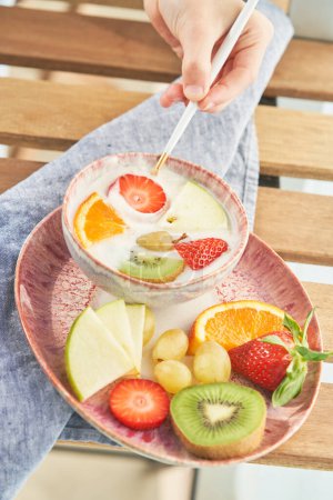 D'en haut de la main de la culture personne anonyme trempant fraise mûre dans un bol avec du yaourt savoureux et divers fruits pendant le petit déjeuner