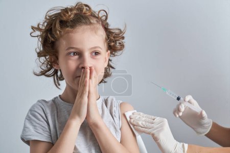 Photo for Crop unrecognizable medic with syringe wiping shoulder of cute boy with folded hands while preparing for injecting in white background - Royalty Free Image