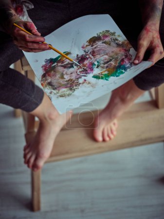 Photo for From above of crop anonymous barefoot kid with dirty hands painting on white paper while sitting on wooden chair in room - Royalty Free Image