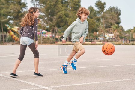 Téléchargez les photos : Délicieux adolescent garçon et fille jouer au basket-ball ensemble tout en passant du temps ensemble en week-end d'été - en image libre de droit