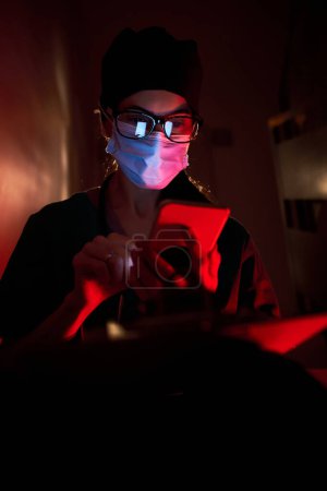 Téléchargez les photos : Femme médecin adulte concernée en uniforme médical et masque de protection assis sur l'escalier et touchant le front tout en naviguant smartphone dans la pièce sombre sous un éclairage rouge - en image libre de droit
