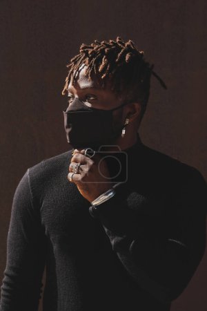 Photo for Concentrated African American male in black shirt and dark protective mask against brown wall looking away - Royalty Free Image